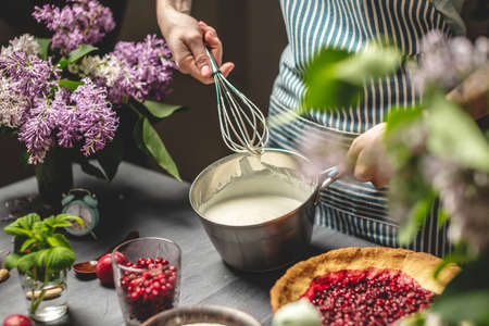 Cooking homemade cranberry pie. A female pastry chef is whipping up a whisk of sour cream. Table with bright ingredients in spring style with flowersの写真素材