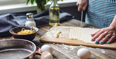 A woman is cutting raw dough with a knife to make homemade noodles. Concept of process of cooking handmade pasta in a cozy atmosphereの写真素材