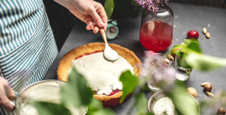 Cooking homemade cranberry pie. A female pastry chef is adding of sour cream with spoon to a baked tart with lingonberries. Table with bright ingredients in spring style with flowersの写真素材