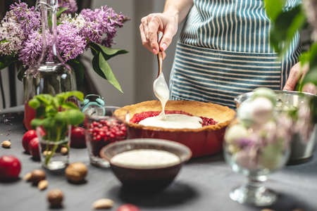 Cooking homemade cranberry pie. A female pastry chef is adding of sour cream with spoon to a baked tart with lingonberries. Table with bright ingredients in spring style with flowersの写真素材
