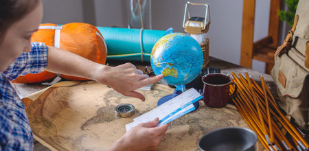 A woman is choosing a place for the next trip on the globe at a wooden table with camping equipment....