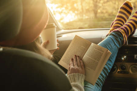 Woman in a car in warm woolen socks yellow is reading a book while sitting in the passenger seat at sunset. Cozy autumn weekend trip. The concept of freedom of travelの写真素材