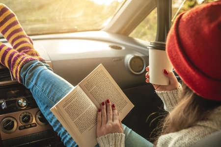 Woman in a car in warm woolen socks yellow is reading a book while sitting in the passenger seat at sunset. Cozy autumn weekend trip. The concept of freedom of travelの写真素材