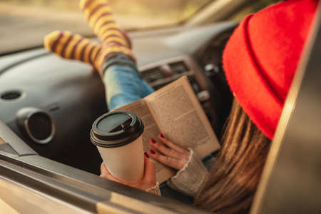 Woman in a car in warm woolen socks yellow is reading a book while sitting in the passenger seat at sunset. Cozy autumn weekend trip. The concept of freedom of travelの写真素材