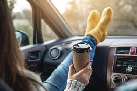 Woman in a car in warm woolen yellow socks is holding a Cup of coffee in hands. Cozy autumn weekend trip. The concept of freedom of travelの写真素材