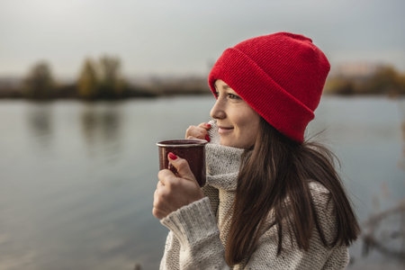 Woman in a red hat is drinking coffee on the lake at sunset. Autumn outdoor recreation. The concept...