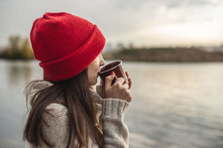 Woman in a red hat is drinking coffee on the lake at sunset. Autumn outdoor recreation. The concept of freedom of travelの写真素材