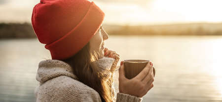 Woman in a red hat is drinking coffee on the lake at sunset. Autumn outdoor recreation. The concept of freedom of travelの写真素材