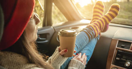 Woman in a car in warm woolen socks yellow is holding a Cup of coffee in hands at sunset. Cozy autumn weekend trip. The concept of freedom of travelの写真素材