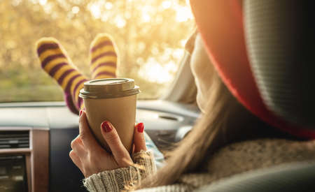 Woman in a car in warm woolen socks yellow is holding a Cup of coffee in hands at sunset. Cozy autumn weekend trip. The concept of freedom of travelの写真素材