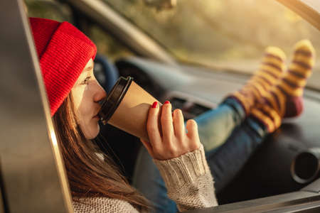 Woman in a car in warm woolen socks yellow is holding a Cup of coffee in hands at sunset. Cozy autumn weekend trip. The concept of freedom of travelの写真素材