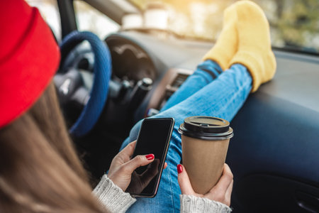 Woman in a car in warm socks is holding a mobile phone gadget and cup of coffee. Cozy autumn weekend...