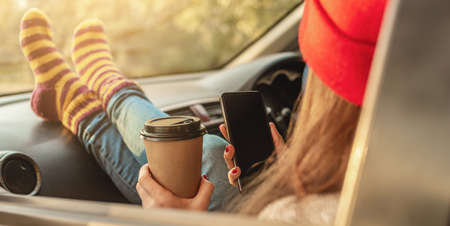 Woman in a car in warm socks is holding a mobile phone gadget and cup of coffee at sunset. Cozy autumn weekend trip. The concept of freedom of travelの写真素材