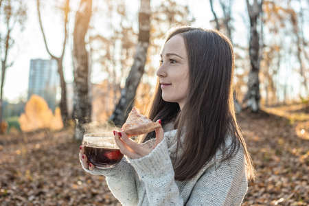 A young pretty woman is eating pizza and drinking hot tea in an autumn yellow Park in the sunset light. The concept of a cozy lifestyle in the autumn cold seasonの写真素材