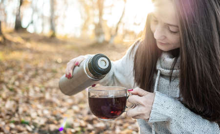 A young pretty woman is pouring hot tea from a thermos in an autumn yellow Park in the light of the sunset. The concept of a cozy lifestyle in the autumn cold seasonの写真素材