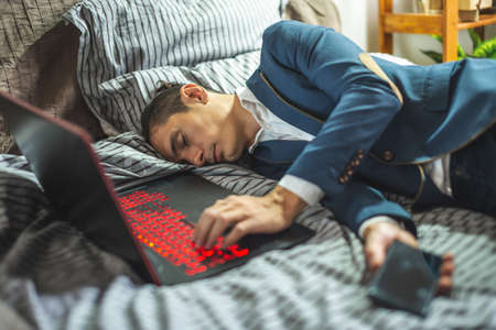 A business man in a suit sleeps in a bed next to a laptop and phone during the day. The concept of fatigue and exhaustion from hard workの写真素材
