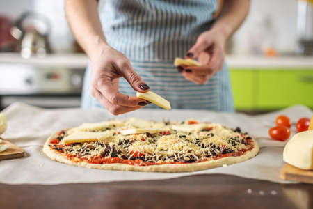 A female chef in an apron is putting mozzarella cheese on a raw pizza. Cooking delicious pizza at home in the kitchen.の写真素材