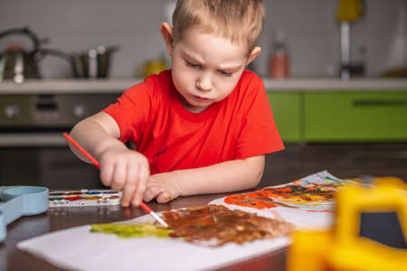 A child in a red T-shirt paints with colorful watercolors at the tableの写真素材