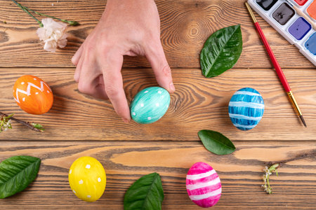Hand is holding a cute green egg painted with colors next to other colorful eggs and fresh spring leaves. Easter and egg decorating conceptの写真素材