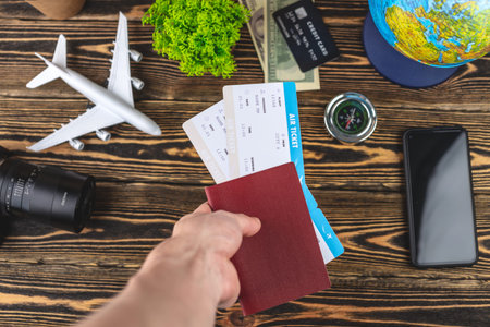 Hand is holding a passport and plane tickets over a wooden table with travel supplies. Vacation and...