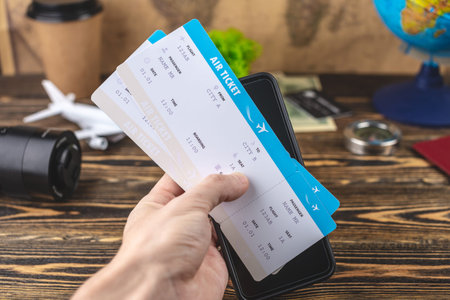 Hand is holding airline tickets and a phone over a wooden table with other travel accessories....