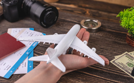 Hand is holding a miniature of plane over a wooden desk with other accessories for a trip. Vacation...
