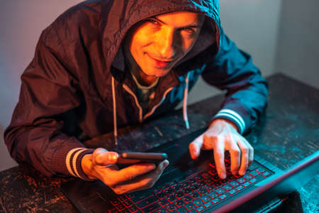 A hacker with a phone is typing on a laptop keyboard in a dark room under a neon light. The concept of cybercrime fraud and identity theftの写真素材
