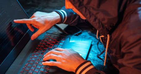 A hooded hacker is typing on a laptop keyboard in a dark room under a neon light. The concept of cybercrime fraud and identity theftの写真素材