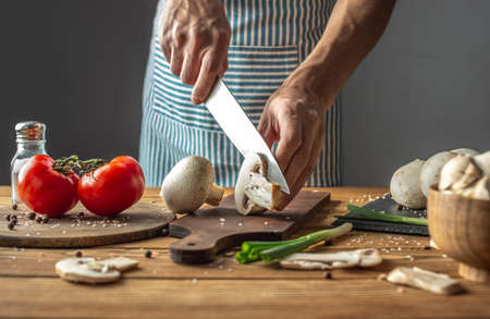 A male chef in a blue apron is cutting fresh mushrooms with a knife. Concept of process of cooking a delicious dish.の写真素材