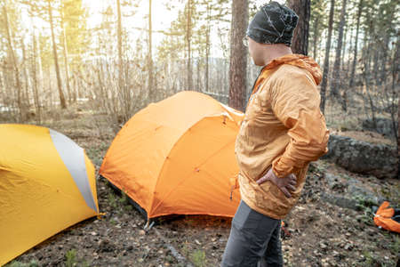 A male traveler is standing in the forest near the tents in a tent camp. Concept of hiking, spending time in nature.の写真素材