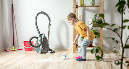 A cute little boy is sweeping the trash from the floor with a broom to a dustpan. Child helps the parents in cleaning the houseの写真素材