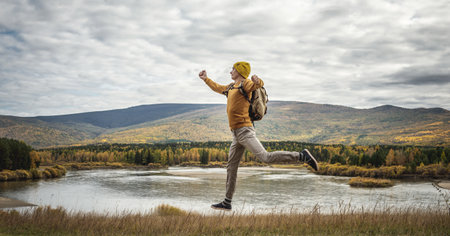 Traveler in a yellow hat and a sweater with a backpack in nature near the river, golden forest and hills is jumping with his arms outstretched. Concept of freedom, travel, hiking and autumn moodの写真素材