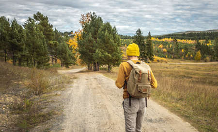 Traveler in a yellow hat and a sweater with a backpack is walking on an empty road along autumn golden forest. Concept of freedom, travel, hiking and autumn moodの写真素材