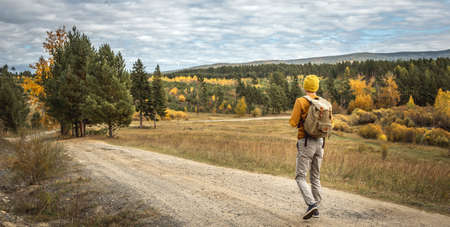 Traveler in a yellow hat and a sweater with a backpack is walking on an empty road along autumn golden forest. Concept of freedom, travel, hiking and autumn moodの写真素材