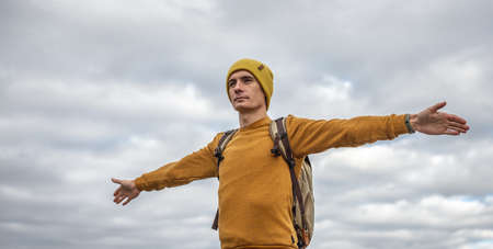 Traveler with a backpack is standing with his arms outstretched to the sides against the background of cloudy autumn sky. Concept of freedom and travelsの写真素材
