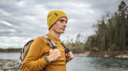 Man traveler in a yellow hat and sweater with a backpack is walking along a fast river with a stony shore. Concept of freedom, travel, hiking and autumn moodの写真素材