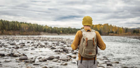 Male traveler is walking along a fast mountain river with huge stones on the shore against the background of an autumn forest. Concept of hiking and travelsの写真素材