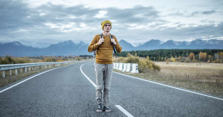 Man traveler with a backpack in a yellow hat and a sweater on an empty asphalt road, against the background of mountains. Concept of freedom, autumn, travel and hikingの写真素材