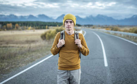 Man traveler with a backpack in a yellow hat and a sweater on an empty asphalt road, in the background of the mountain. Concept of autumn mood, travel and hikingの写真素材