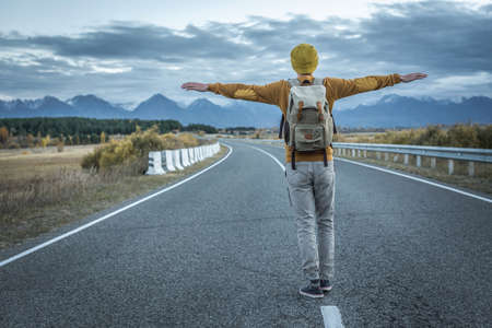 Traveler with a backpack in a yellow hat and a sweater on a road, against the background of mountains is standing with his arms outstretched to the sides. Concept of freedom, autumn, travel, hikingの写真素材