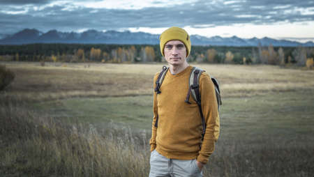 Traveler with a backpack in a yellow hat and a sweater in nature against the background of an autumn golden forest and mountains. Concept of travel, hiking and autumn moodの写真素材