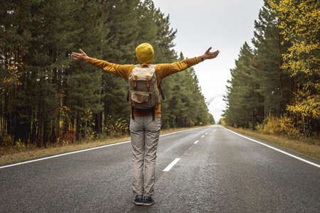 Man in a yellow hat and a sweater on a smooth empty asphalt road along a beautiful golden forest is standing with his arms outstretched to the sides. Concept of freedom, autumn, travel, hikingの写真素材