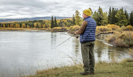 Male fisherman is standing on the river bank in the golden autumn forest with a fishing rod in his hands and catching fish. Concept of calm outdoor recreation, pastime, hobbyの写真素材