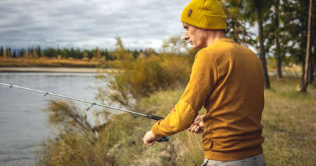 Young man fisherman in a yellow sweater and hat is fishing on the river bank in the autumn forest with a spinning rod in his hands. Concept of relaxing outdoor recreation, lifestyle, hobbyの写真素材