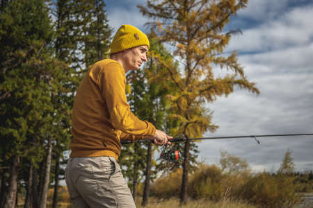 Young man fisherman in a yellow sweater and hat is fishing on the river bank in the autumn forest with a spinning rod in his hands. Concept of relaxing outdoor recreation, lifestyle, hobbyの写真素材