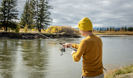 Young man fisherman in a yellow sweater and hat is fishing on the river bank in the autumn forest with a spinning rod in his hands. Concept of relaxing outdoor recreation, lifestyle, hobbyの写真素材