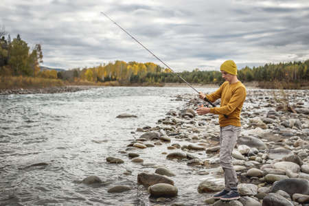 Young man fisherman in a yellow sweater and hat is fishing on the stony river bank in the autumn forest with a spinning rod in his hands. Concept of relaxing outdoor recreation, lifestyle, hobbyの写真素材