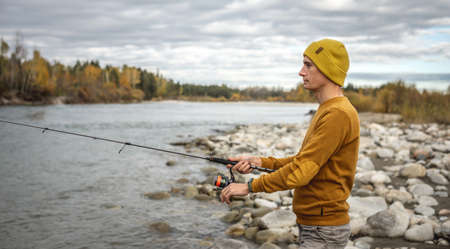 Young man fisherman in a yellow sweater and hat is fishing on the stony river bank in the autumn forest with a spinning rod in his hands. Concept of relaxing outdoor recreation, lifestyle, hobbyの写真素材