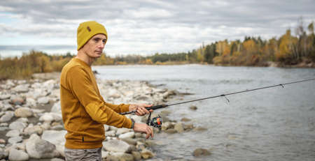Young man fisherman in a yellow sweater and hat is fishing on the stony river bank in the autumn forest with a spinning rod in his hands. Concept of relaxing outdoor recreation, lifestyle, hobbyの写真素材