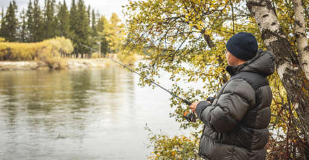 Male fisherman is standing on the river bank in the golden autumn forest with a fishing rod in his hands and catching fish. Concept of calm outdoor recreation, pastime, hobbyの写真素材
